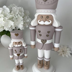 May include: Two nutcracker figurines in a muted mauve, white, and tan color palette. Each figure features a tall hat with a heart emblem, a white mustache, and heart-shaped buttons on the jacket and boots, all standing on a heart-shaped base.