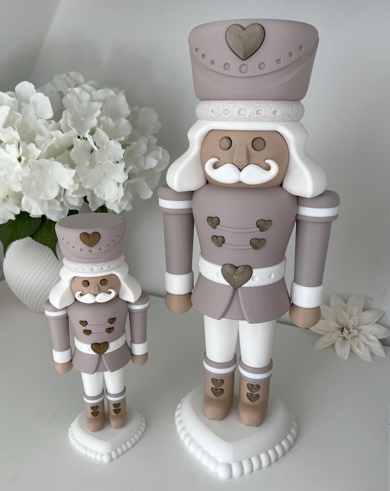 May include: Two nutcracker figurines in a muted mauve, white, and tan color palette. Each figure features a tall hat with a heart emblem, a white mustache, and heart-shaped buttons on the jacket and boots, all standing on a heart-shaped base.