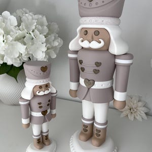 May include: Two nutcracker figurines in a taupe, white, and tan color palette. Each figure features a tall hat with a heart emblem, a white mustache, and heart-shaped buttons on the jacket and boots. They stand on white heart-shaped bases.