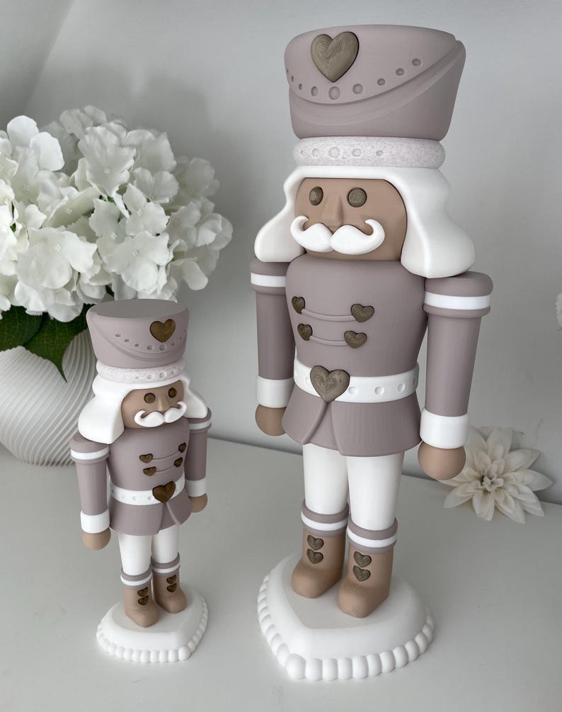 May include: Two nutcracker figurines in a taupe, white, and tan color palette. Each figure features a tall hat with a heart emblem, a white mustache, and heart-shaped buttons on the jacket and boots. They stand on white heart-shaped bases.