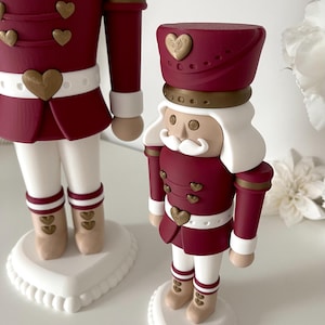 May include: Two nutcracker figures in a burgundy and white color scheme. Each figure wears a burgundy jacket with gold heart-shaped buttons, a white belt, white trousers, and tan boots. They stand on white heart-shaped bases with white hair.