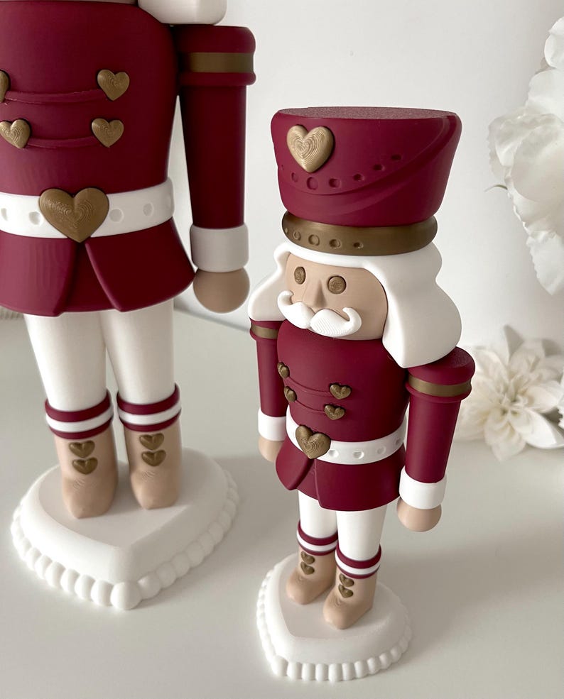 May include: Two nutcracker figures in a burgundy and white color scheme. Each figure wears a burgundy jacket with gold heart-shaped buttons, a white belt, white trousers, and tan boots. They stand on white heart-shaped bases with white hair.