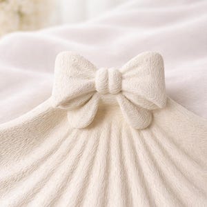 May include: A cream-colored decorative bow, possibly ceramic, sits atop a textured, fan-shaped surface. The bow has a detailed, carved design, and the surface has a ribbed pattern. The background is a soft, blurred white.