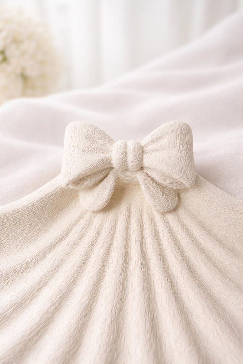 May include: A cream-colored decorative bow, possibly ceramic, sits atop a textured, fan-shaped surface. The bow has a detailed, carved design, and the surface has a ribbed pattern. The background is a soft, blurred white.