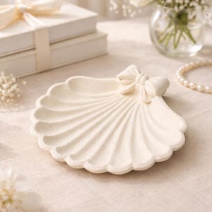May include: Off-white seashell-shaped decorative dish with a bow accent. The dish has a textured surface and scalloped edges. The background includes white roses, a pearl necklace, and a stack of white boxes tied with a ribbon.