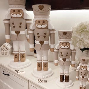May include: Four nutcracker figures in varying heights of 70cm, 58cm, 40cm, and 24cm. Each figure features a beige and white color scheme with brown heart-shaped accents on the hat, jacket, and boots, and a white mustache and hair.