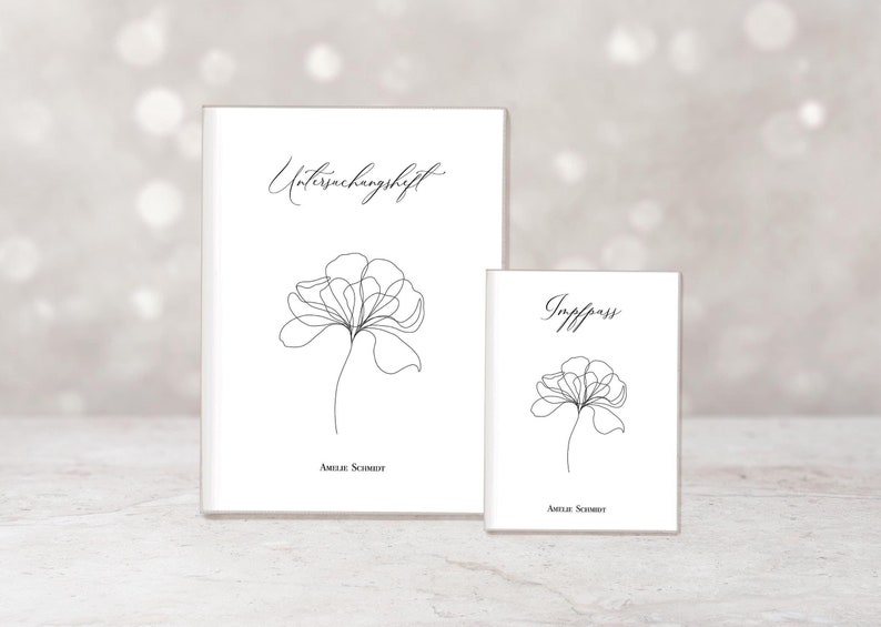 Op de afbeelding: Twee witte notitieboekjes met zwarte bloemmotieven. Het grotere notitieboekje heeft het woord "Untersuchungheft" en het kleinere het woord "Impfpass". Beide notitieboekjes hebben de naam "Amelie Schmidt" onderaan.