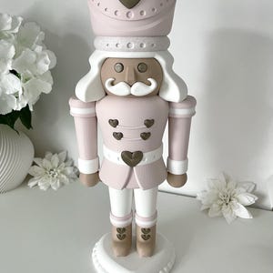 May include: A decorative nutcracker figure in a soft pink, white, and metallic bronze color palette. The figure features a tall hat with a bronze heart, a white mustache, and bronze heart-shaped buttons on the jacket and boots, standing on a heart-shaped base.