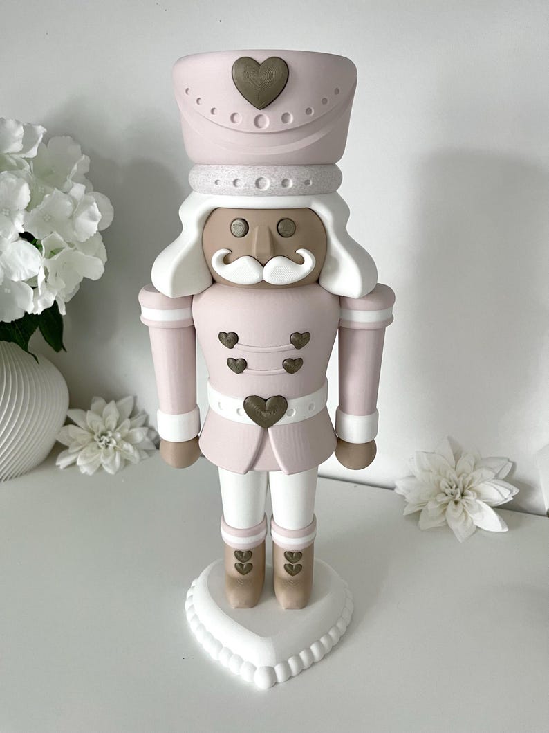 May include: A decorative nutcracker figure in a soft pink, white, and metallic bronze color palette. The figure features a tall hat with a bronze heart, a white mustache, and bronze heart-shaped buttons on the jacket and boots, standing on a heart-shaped base.