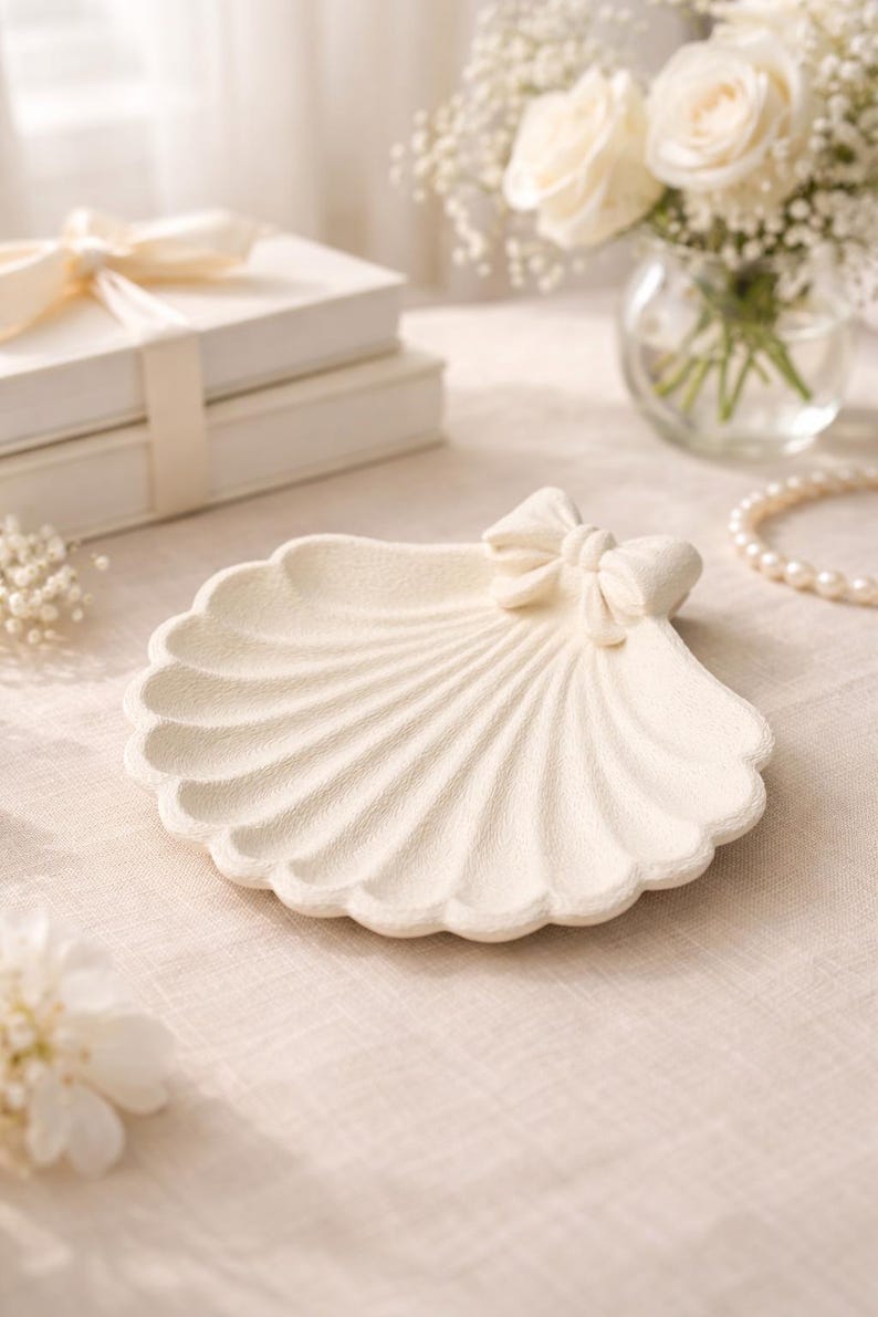 May include: Off-white seashell-shaped decorative dish with a bow accent. The dish has a textured surface and scalloped edges. The background includes white roses, a pearl necklace, and a stack of white boxes.