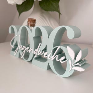 May include: A mint green decorative sign with the numbers "2026" and the word "Jugendweihe" in white script. A small glass bottle with a cork stopper sits between the numbers. White floral accents are on the right side.