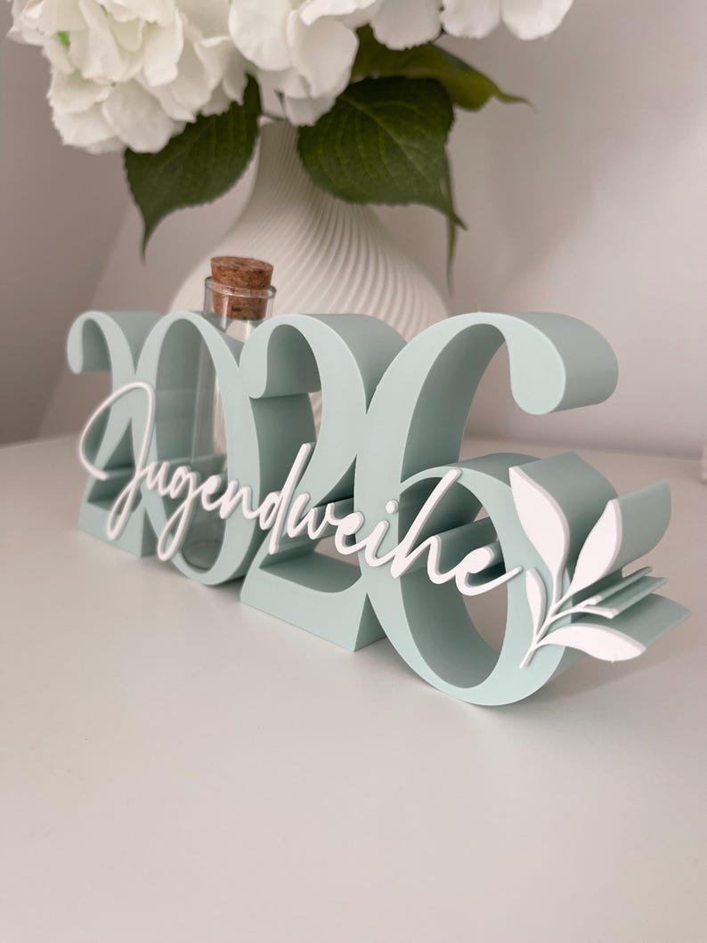 May include: A mint green decorative sign with the numbers "2026" and the word "Jugendweihe" in white script. A small glass bottle with a cork stopper sits between the numbers. White floral accents are on the right side.