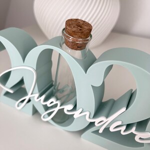 May include: A light teal-colored decorative piece with the word "Jugendweihe" in white script. A small glass bottle with a cork stopper is nestled within the design. A white, textured vase is in the background.