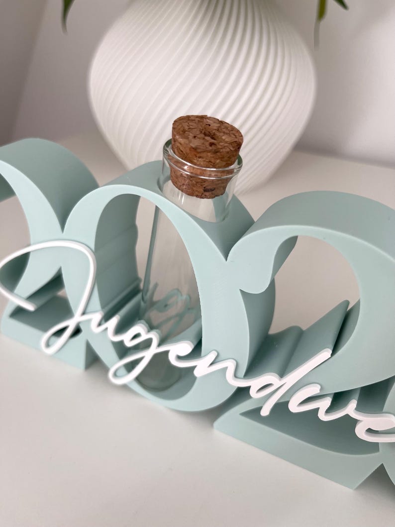 May include: A light teal-colored decorative piece with the word "Jugendweihe" in white script. A small glass bottle with a cork stopper is nestled within the design. A white, textured vase is in the background.