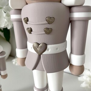 May include: A nutcracker figurine in a muted taupe and white color palette. The figure features a white mustache, a taupe jacket with four small bronze heart-shaped buttons, and a white belt with a large central bronze heart-shaped buckle.