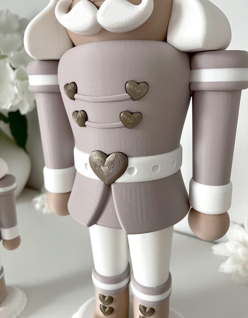 May include: A nutcracker figurine in a muted taupe and white color palette. The figure features a white mustache, a taupe jacket with four small bronze heart-shaped buttons, and a white belt with a large central bronze heart-shaped buckle.