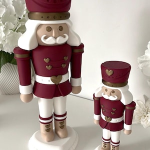 May include: Two nutcracker figurines in varying sizes, featuring a deep burgundy uniform with gold heart-shaped buttons and accents. Each figure has a white mustache, white hair, and stands on a white circular base with a heart-shaped platform.