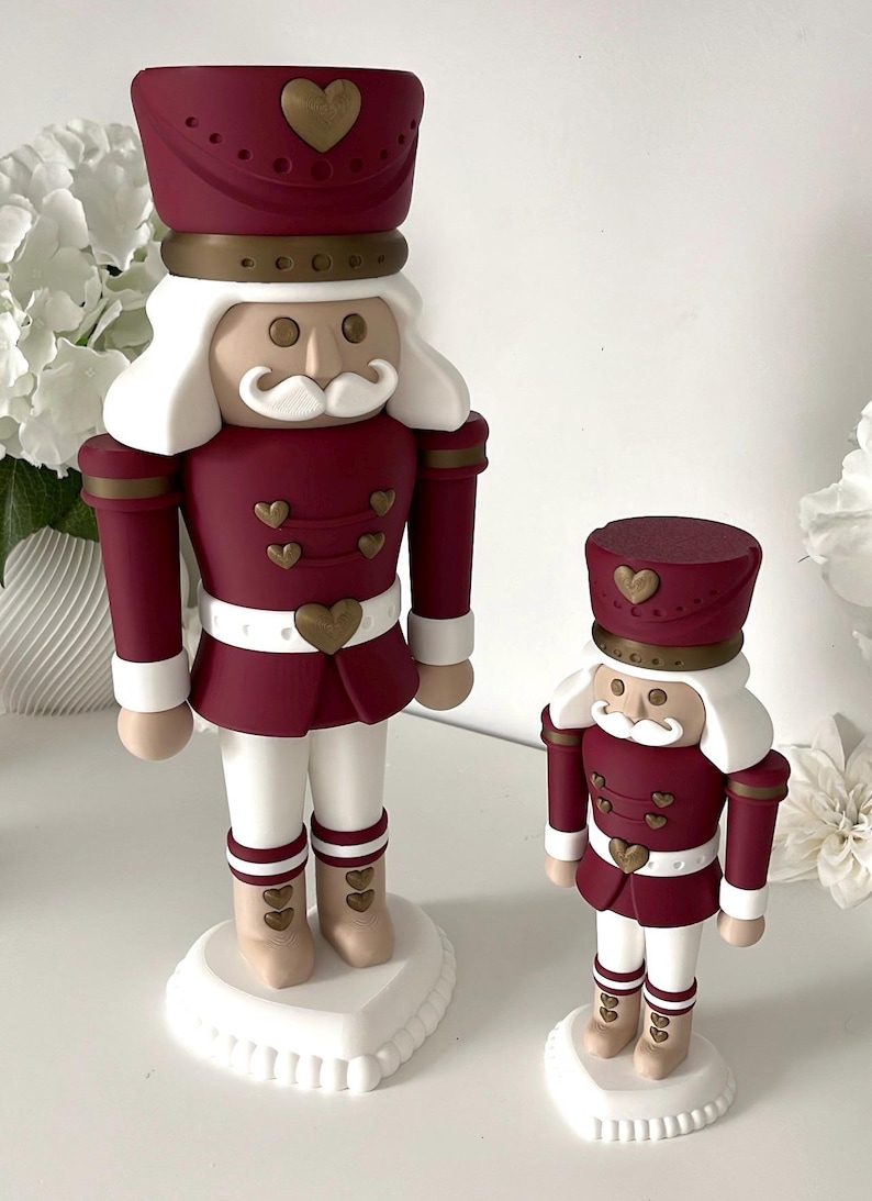 May include: Two nutcracker figurines in varying sizes, featuring a deep burgundy uniform with gold heart-shaped buttons and accents. Each figure has a white mustache, white hair, and stands on a white circular base with a heart-shaped platform.