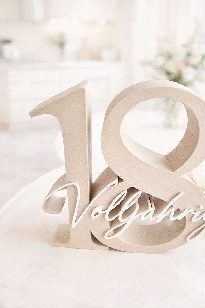May include: A wooden number 18 cake topper in a light beige color. The number 18 is stylized with a cursive word, "Vollj&auml;hrig", layered across the bottom. The cake topper is on a white surface.