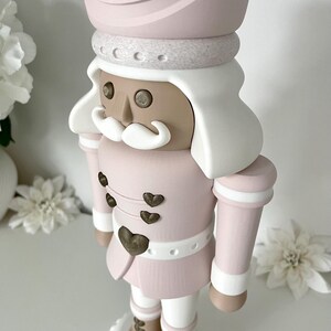 May include: A decorative nutcracker figure in a soft pastel pink, white, and tan color palette. The figure wears a tall pink hat with a heart emblem, features a white mustache, and has small heart-shaped buttons on the jacket front.