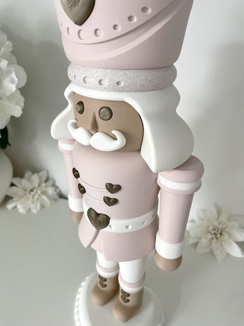 May include: A decorative nutcracker figure in a soft pastel pink, white, and tan color palette. The figure wears a tall pink hat with a heart emblem, features a white mustache, and has small heart-shaped buttons on the jacket front.