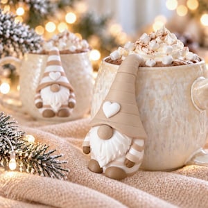 Könnte beinhalten: Zwei beige gesprenkelte Keramiktassen gefüllt mit heißer Schokolade, garniert mit Schlagsahne und Mini-Marshmallows. Jede Tasse zeigt eine modellierte Zwergenfigur mit einem beige gestreiften Hut, einem weißen Herzakzent und einem langen weißen Bart.