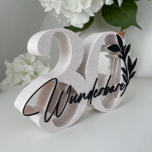 May include: A freestanding number 30 decoration with a speckled light beige finish. The number features black cursive text reading "Wunderbare" across the front, accented by a black leaf design and a small glass tube insert with a cork stopper.