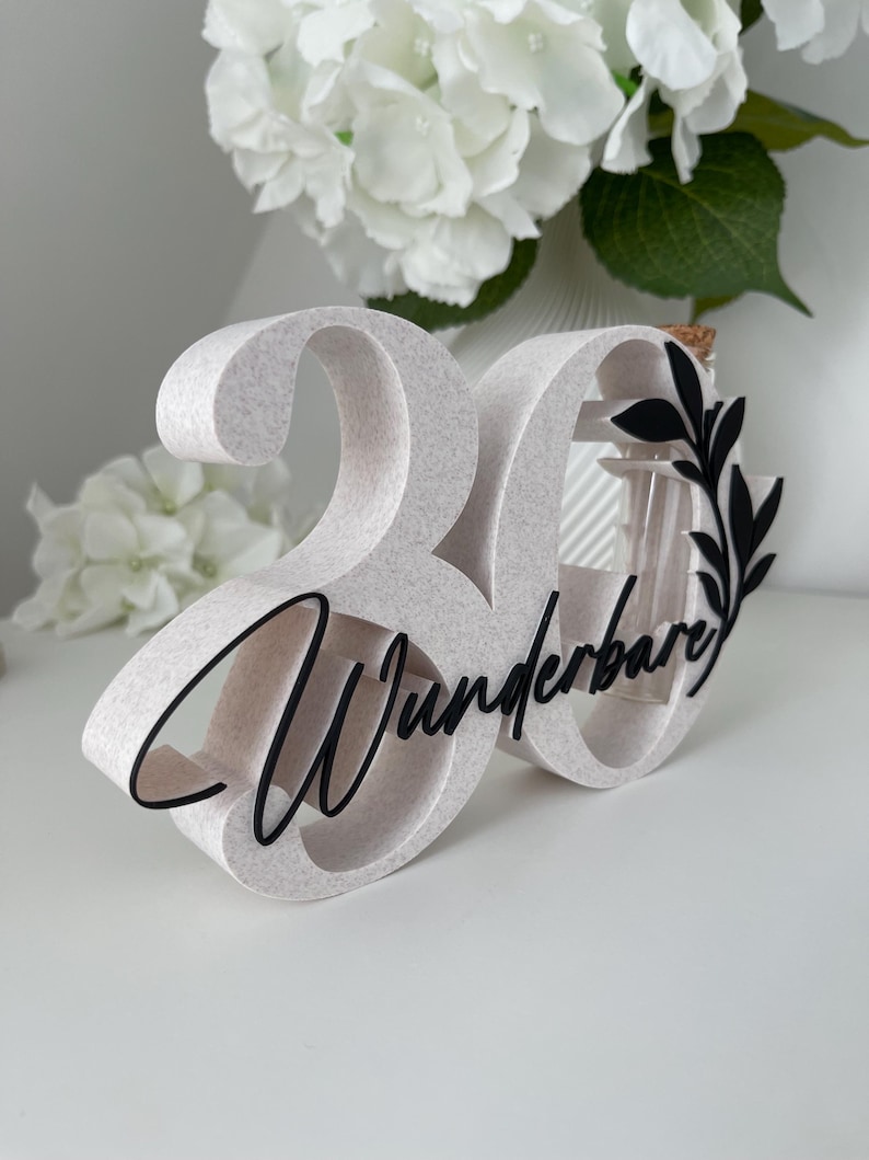 May include: A freestanding number 30 decoration with a speckled light beige finish. The number features black cursive text reading "Wunderbare" across the front, accented by a black leaf design and a small glass tube insert with a cork stopper.