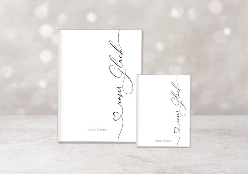 May include: Two white rectangular objects, one large and one small, with the words "unser Gl&uuml;ck" in elegant black script. A heart shape is incorporated into the script. The name "Amelie Schmidt" is printed at the bottom.
