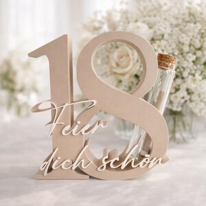 May include: A beige wooden number 18 with the words "Feier dich sch&ouml;n" in white script. A small glass vial with a cork stopper is inserted into the number 8. The background features white flowers.