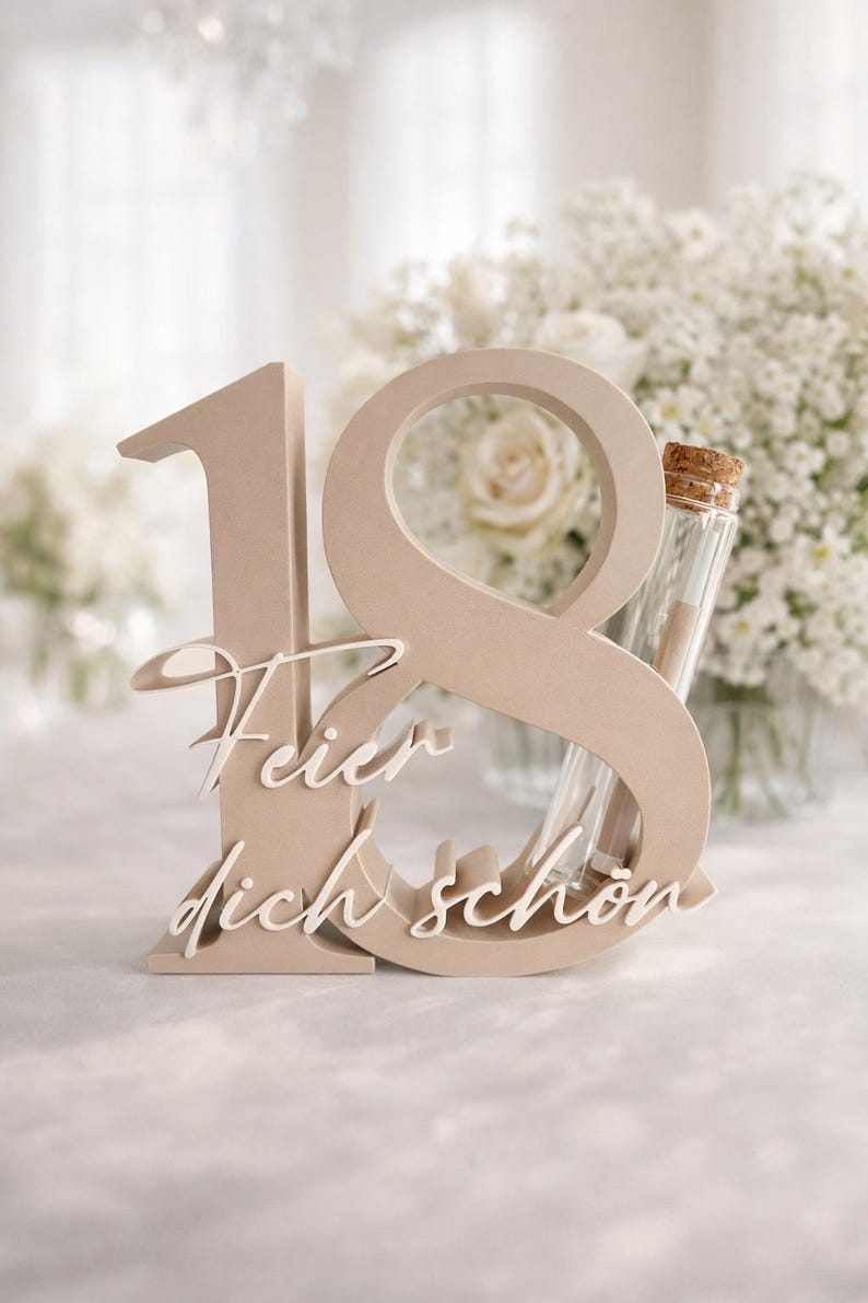 May include: A beige wooden number 18 with the words "Feier dich sch&ouml;n" in white script. A small glass vial with a cork stopper is inserted into the number 8. The background features white flowers.