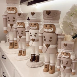 May include: A collection of five nutcracker figurines in varying sizes, featuring a neutral color palette of beige, white, and metallic bronze. Each nutcracker is adorned with small heart-shaped accents on the hat, jacket, and base.