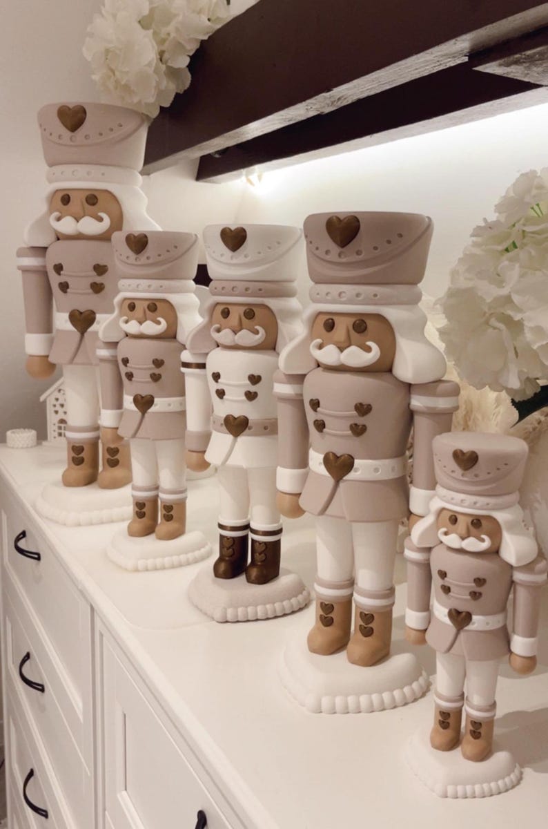 May include: A collection of five nutcracker figurines in varying sizes, featuring a neutral color palette of beige, white, and metallic bronze. Each nutcracker is adorned with small heart-shaped accents on the hat, jacket, and base.