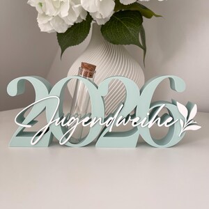 May include: A mint green wooden decoration with the numbers "2026" and the word "Jugendweihe" in white script. A small glass vial with a cork stopper is centered within the numbers. White floral arrangement in a white vase is in the background.