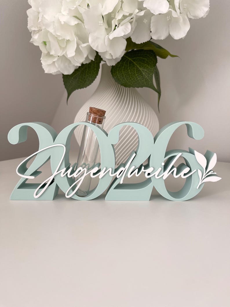 May include: A mint green wooden decoration with the numbers "2026" and the word "Jugendweihe" in white script. A small glass vial with a cork stopper is centered within the numbers. White floral arrangement in a white vase is in the background.