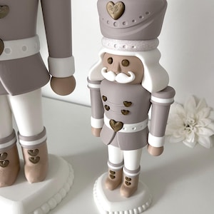 May include: A nutcracker figurine in shades of taupe, white, and metallic gold. The figure wears a taupe uniform with gold heart-shaped buttons, a white mustache, and a hat adorned with a gold heart. It stands on a white heart-shaped base.