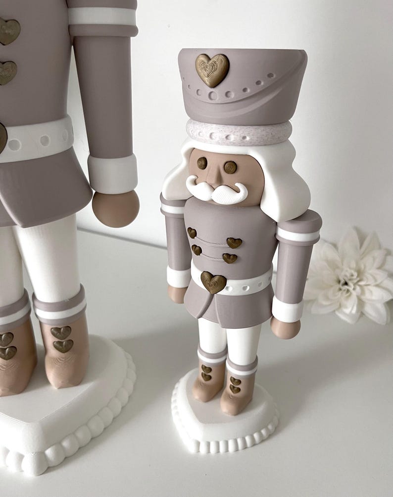 May include: A nutcracker figurine in shades of taupe, white, and metallic gold. The figure wears a taupe uniform with gold heart-shaped buttons, a white mustache, and a hat adorned with a gold heart. It stands on a white heart-shaped base.