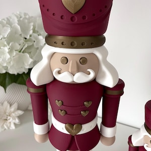 May include: A nutcracker figurine featuring a deep burgundy uniform with gold heart-shaped buttons and accents. The figure has white hair and a mustache, a tall burgundy hat with a gold heart, and white trousers. The design includes gold circular details.
