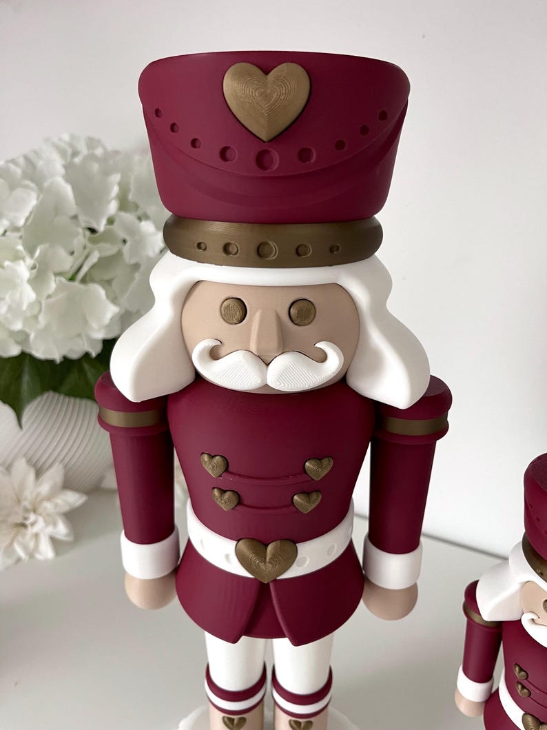 May include: A nutcracker figurine featuring a deep burgundy uniform with gold heart-shaped buttons and accents. The figure has white hair and a mustache, a tall burgundy hat with a gold heart, and white trousers. The design includes gold circular details.