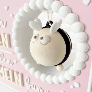 May include: A pink rectangular wall plaque featuring a white circular frame with a hanging 3D bee figure. The bee has a cream-coloured body, black stripes, and heart-shaped wings. The plaque displays white raised text including the word bummeln.