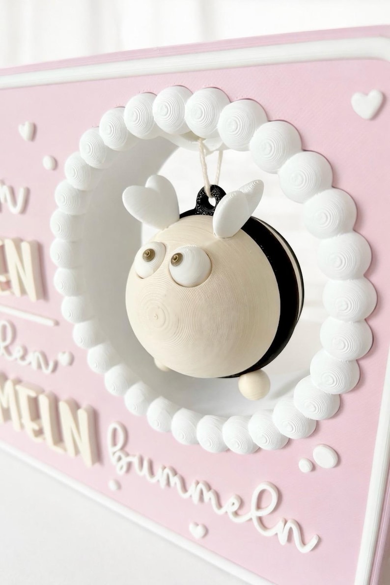 May include: A pink rectangular wall plaque featuring a white circular frame with a hanging 3D bee figure. The bee has a cream-coloured body, black stripes, and heart-shaped wings. The plaque displays white raised text including the word bummeln.