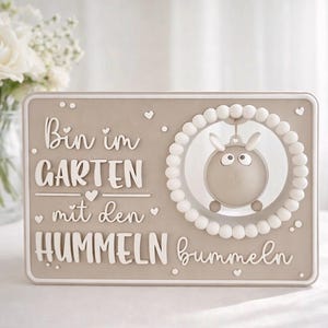 May include: A beige rectangular plaque with white raised lettering and a cartoon sheep design. The text reads "Bin im GARTEN mit den HUMMELN bummeln". The sheep is inside a white beaded circle, and small hearts are scattered around the edges.