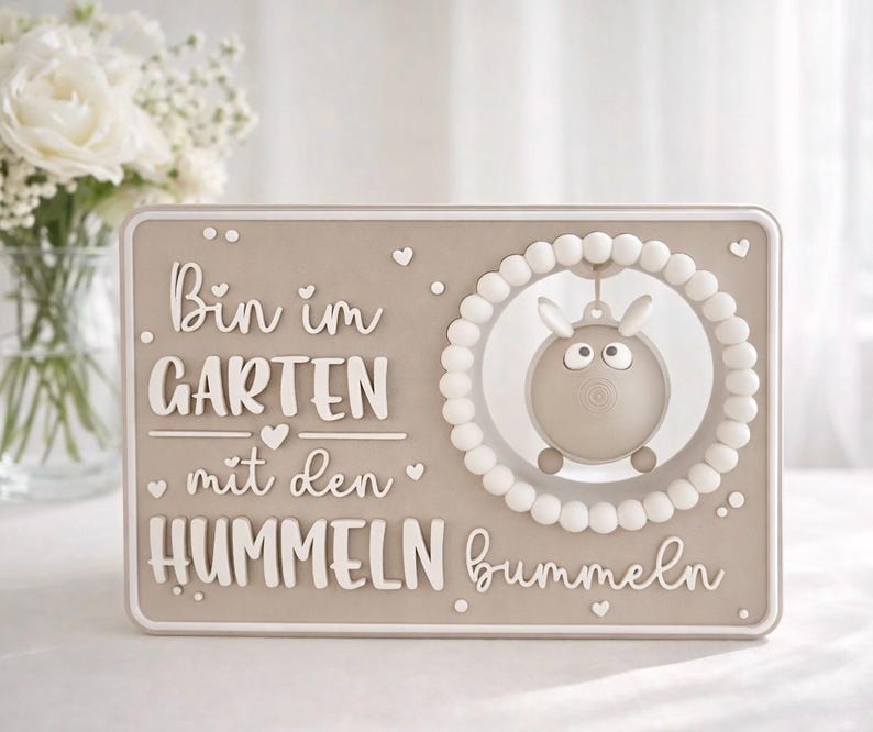 May include: A beige rectangular plaque with white raised lettering and a cartoon sheep design. The text reads "Bin im GARTEN mit den HUMMELN bummeln". The sheep is inside a white beaded circle, and small hearts are scattered around the edges.