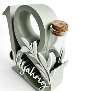 May include: A sage green decorative piece with the number 19, a small glass bottle with a cork, and white leaf accents. The word "Vollj&auml;hrig" is written in cursive. The design is on a white background.