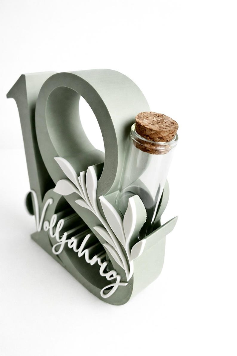 May include: A sage green decorative piece with the number 19, a small glass bottle with a cork, and white leaf accents. The word "Vollj&auml;hrig" is written in cursive. The design is on a white background.