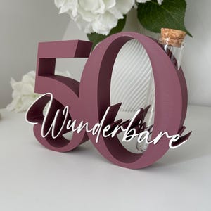 May include: A dark mauve wooden number 50 sign with the white cursive text Wunderbare attached to the front. A clear glass test tube with a cork stopper is inserted into the top of the number zero, serving as a small vase for floral stems.
