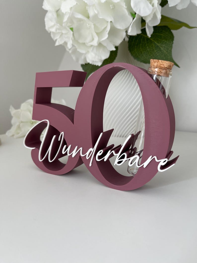 May include: A dark mauve wooden number 50 sign with the white cursive text Wunderbare attached to the front. A clear glass test tube with a cork stopper is inserted into the top of the number zero, serving as a small vase for floral stems.