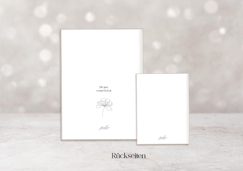 May include: Two white rectangular cards, one large and one small, are displayed against a soft, blurred background. The larger card features the text "Du bist unser Gl&uuml;ck" and a delicate floral illustration. The word "R&uuml;ckseiten" is at the bottom.