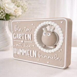 May include: A beige rectangular plaque with white lettering and a 3D cartoon bee. The text reads "Bin im GARTEN mit den HUMMELN bummeln". The bee is framed by a white beaded circle. Decorative item.