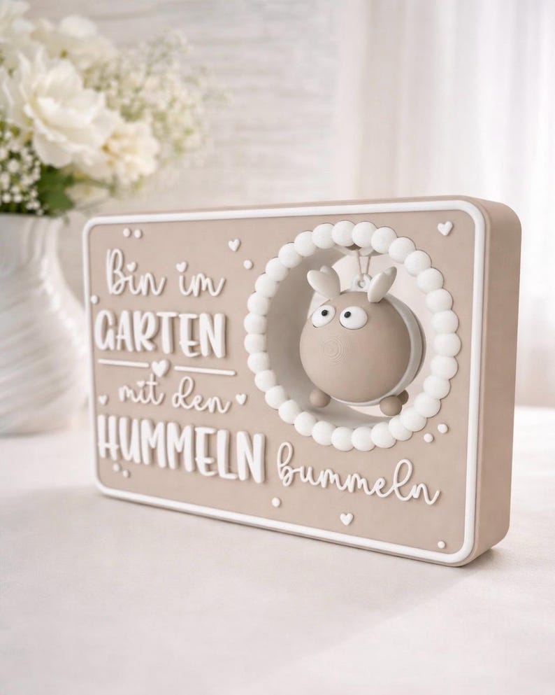 May include: A beige rectangular plaque with white lettering and a 3D cartoon bee. The text reads "Bin im GARTEN mit den HUMMELN bummeln". The bee is framed by a white beaded circle. Decorative item.